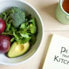 Style: natural, slightly humorous, with a "clean and earthy" aesthetic. Scene: a kitchen counter with a bowl of colorful vegetables – beets, broccoli, arugula, avocado, garlic. Next to it, a mug of herbal tea and a notebook with the words "Detox from the Kitchen" written on it. Soft, natural lighting, pastel colors (green, burgundy, beige). Mood: freshness, lightness, a sense of renewal.
